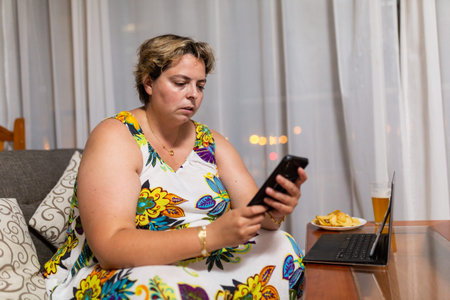 woman sitting on the sofa with cell phone in hand, laptop on the table with foodの写真素材