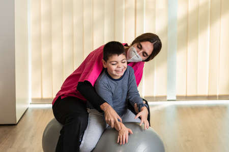 disabled child and physiotherapist on top of a Peanut Gym Ball doing balance exercises. pandemic mask protectionの写真素材
