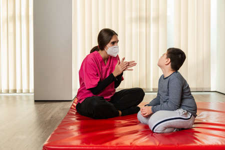 disabled child and physiotherapist on a red gymnastic mat doing exercises. pandemic mask protectionの写真素材