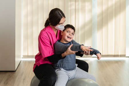 disabled child and physiotherapist on top of a Peanut Gym Ball doing balance exercises. pandemic mask protectionの写真素材