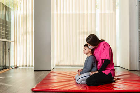 disabled child and physiotherapist on a red gymnastic mat doing exercises. pandemic mask protectionの写真素材