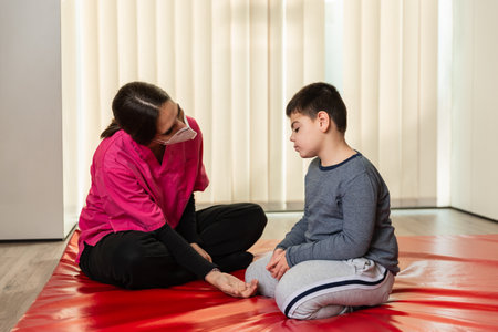 disabled child and physiotherapist on a red gymnastic mat doing exercises. pandemic mask protectionの写真素材