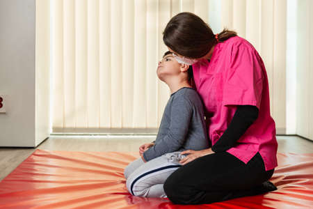 disabled child and physiotherapist on a red gymnastic mat doing exercises. pandemic mask protectionの写真素材