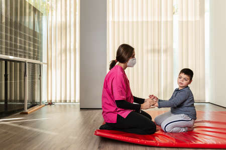 disabled child and physiotherapist on a red gymnastic mat doing exercises. pandemic mask protectionの写真素材