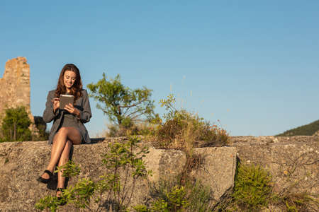 Young female remote employee in black formal suit using tablet, while working online sitting on stone in hilly terrain against cloudless blue sky in summer dayの写真素材