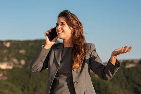 Positive adult businesswoman in formal gray suit answering phone call and looking to the side, while standing against blurred green hills and blue sky in sunny summer dayの写真素材