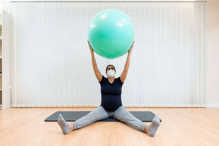 Close up of a pregnant woman with face mask sitting on floor doing pilates exercises with a ballの写真素材
