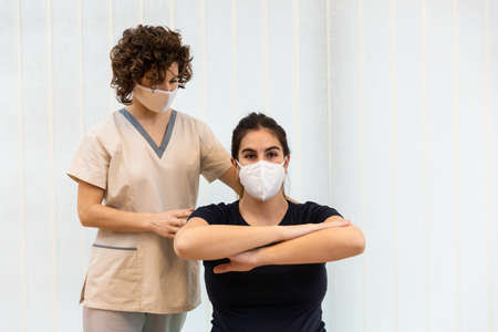 A pregnant woman wearing a face mask doing exercises with her arms in front of her with the help of physiotherapist.の写真素材