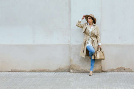 An adult woman with a hat and purse leaning against a wall while looking awayの写真素材