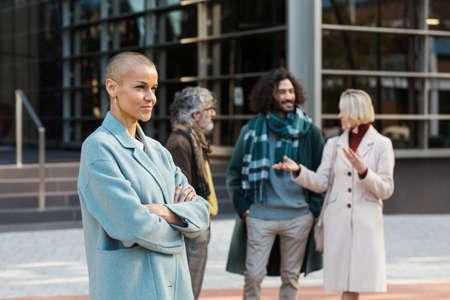 Professional businesswoman standing with arms crossed outdoors while colleagues talking in the background.の写真素材