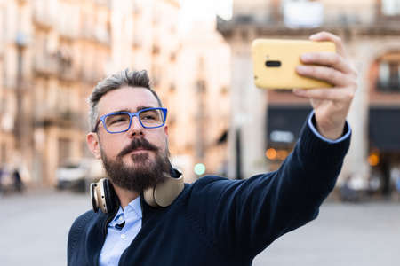 Man with sunglasses and wireless headphones taking a selfie with a mobile phone outdoors on the street.の写真素材