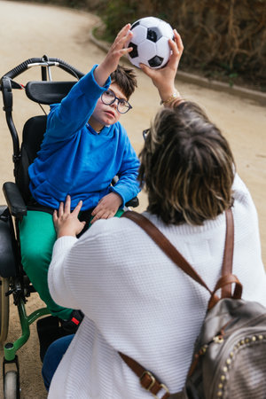 A mother playing with a soccer ball with her son with a disability in a wheelchair in the park.の写真素材