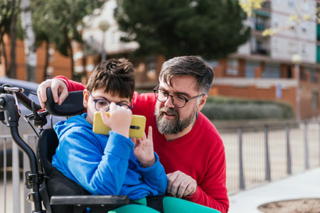 Disabled child in wheelchair watching something on the mobile phone with his father.の写真素材