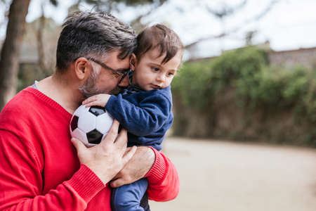 Man holds his little son and a soccer ball while playing together outdoors in the park.の写真素材