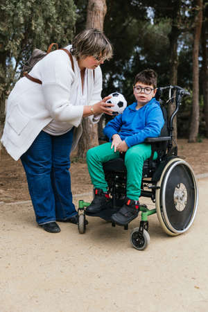 A mother giving a soccer ball to her son with a disability who uses a wheelchair in the park to play