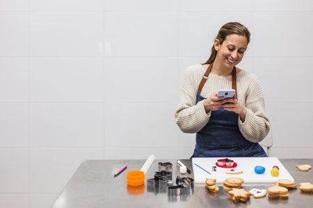 Smiling pastry chef using her mobile phone while working in the kitchen of the pastry shop.の写真素材