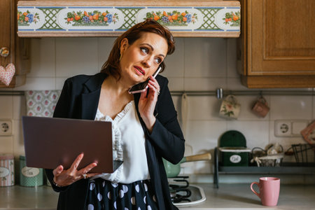 Businesswoman talking on the phone while using laptop standing in the kitchen at home.の写真素材