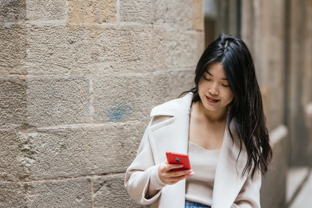 Asian woman smiling while using her mobile phone outdoors.の写真素材