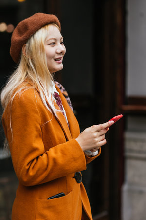 Stylish woman smiling while holding mobile phone outdoors.の写真素材