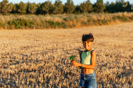 Cheerful young girl in casual clothes holds a potted plant in the field in summer.の写真素材
