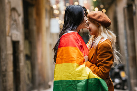 Woman wrapped in lgbt rainbow flag while kissing her girlfriend on the forehead.の写真素材