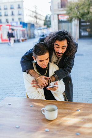 Gay couple hugging while looking at the mobile phone outdoors at a cafe.の写真素材