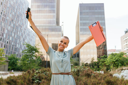A businesswoman with arms raised in a triumphant gesture, holding a phone and a folder.の写真素材
