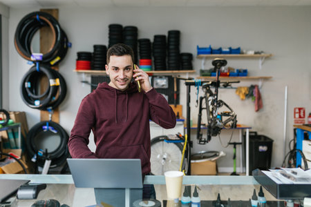 The bike mechanic is answering the phone with a smile, looking at the camera with a laptop in front of him on the counter of his bike workshop.の写真素材