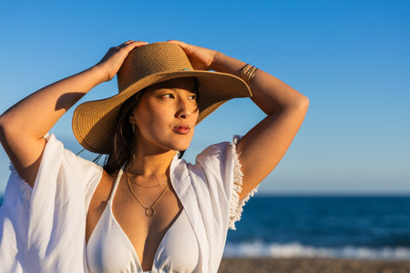 Young woman in a stylish hat enjoys the serene beach atmosphere, embodying the relaxed essence of summer by the seaの写真素材
