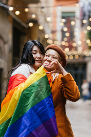 Lesbian couple making heart shape with hands while holding the rainbow flag outdoors.の写真素材