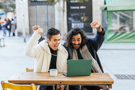 Two male friends celebrating success while watching football match on laptop in a cafeの写真素材