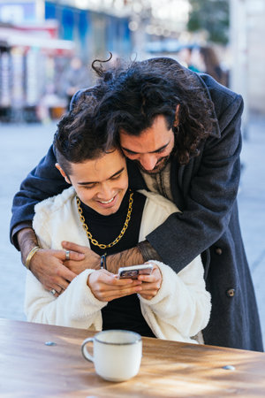 Young man embracing his boyfriend using smartphone at a cafeの写真素材