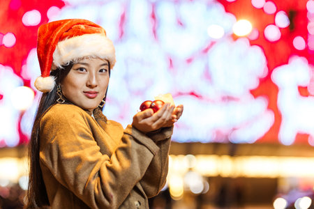 Young woman wearing santa hat holding christmas baubles in city at nightの写真素材