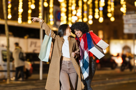 Two happy young women holding shopping bags and walking in a christmas street at nightの写真素材
