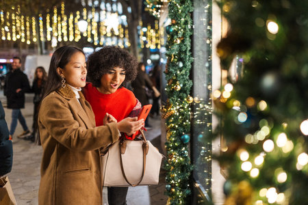 Two young women window shopping while checking smartphone in a decorated city at christmas timeの写真素材