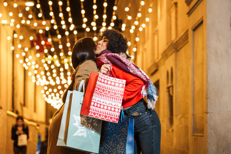 Two women are hugging and holding shopping bags on a decorated street at christmas timeの写真素材