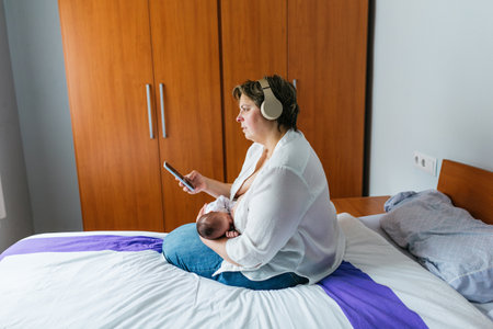 Mother breastfeeding newborn baby and using smartphone while wearing headphonesの写真素材