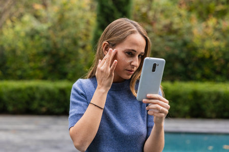 Woman applying cream while looking at smartphone outdoorsの写真素材