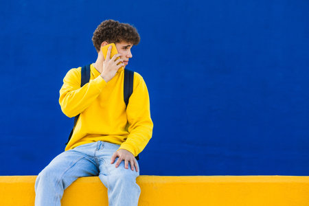 Young man with backpack using smartphone sitting on yellow wall with blue backgroundの写真素材