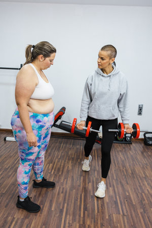 Personal trainer showing correct biceps curl form to woman in gymの写真素材