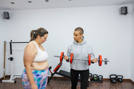 Personal trainer showing correct biceps curl form to overweight woman in gymの写真素材