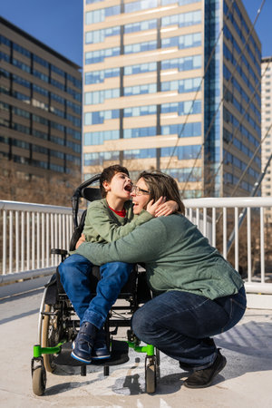 Mother hugging her son with disability with cerebral palsy in a wheelchair on a bridgeの写真素材
