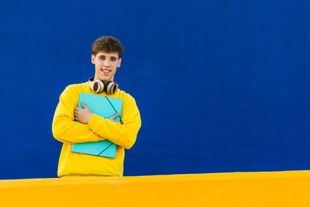 Smiling college student holding a binder and wearing headphones on a blue and yellow backgroundの写真素材