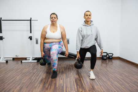 Personal trainer supporting overweight client doing kettlebell lunges in gymの写真素材