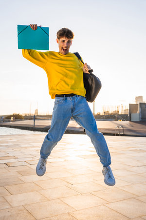 Excited student jumping with binder and backpack at sunsetの写真素材