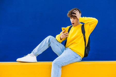 Shocked college student reacting to news on smartphone while sitting on yellow wall