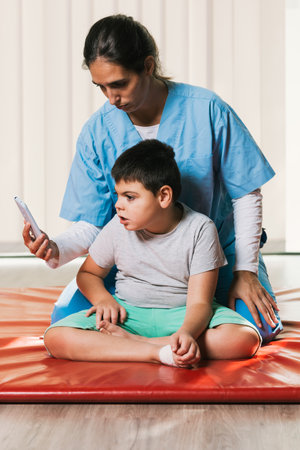 Physiotherapist showing smartphone to child with cerebral palsy during rehabilitation sessionの写真素材