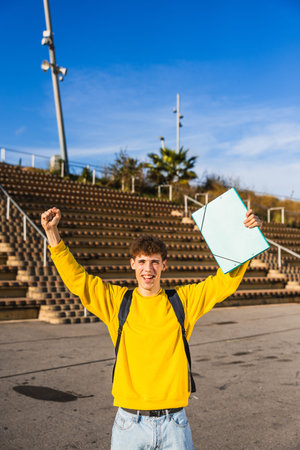 Happy college student celebrating with raised arms and folder in university campusの写真素材