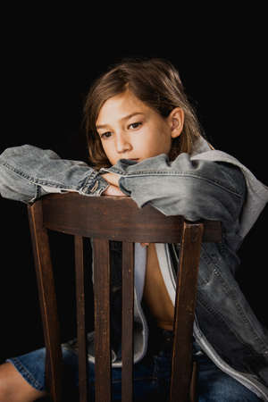 Eleven years old boy with hooded sweater and a jean jacket sitting on a wood chair against a black backgroundの写真素材