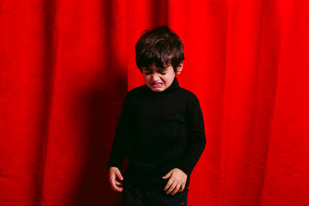 Three years old boy, wearing black clothes, crying against a red curtainの写真素材